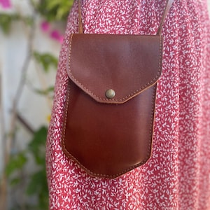 May include: A brown leather crossbody bag with a flap closure and a single button. The bag is worn over a red floral dress.