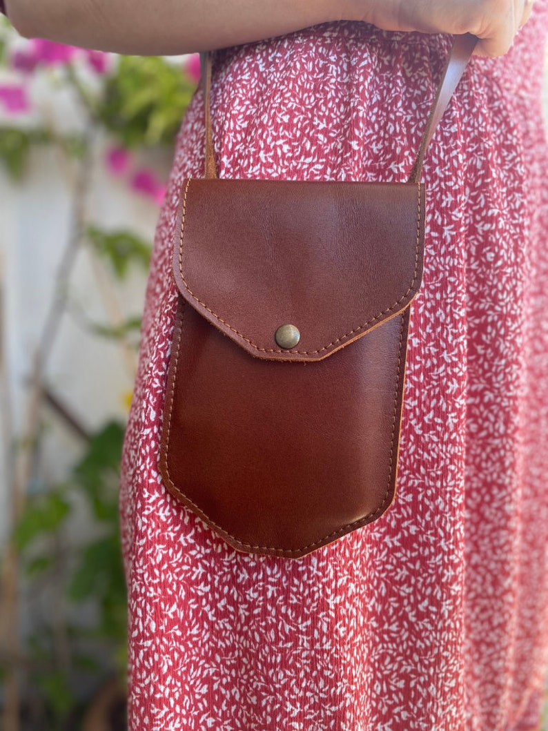 May include: A brown leather crossbody bag with a flap closure and a single button. The bag is worn over a red floral dress.