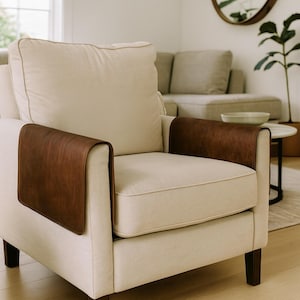 May include: Cream armchair with dark brown leather arm covers. The chair has a square back and armrests, with a light-coloured welt cord trim. The legs are dark brown. The chair is in a well-lit room.