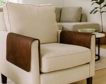 May include: An off-white upholstered armchair featuring two rectangular dark brown leather armrest covers draped over the sides. The chair has a thick back cushion and a seat cushion with piping detail, supported by dark wooden legs.