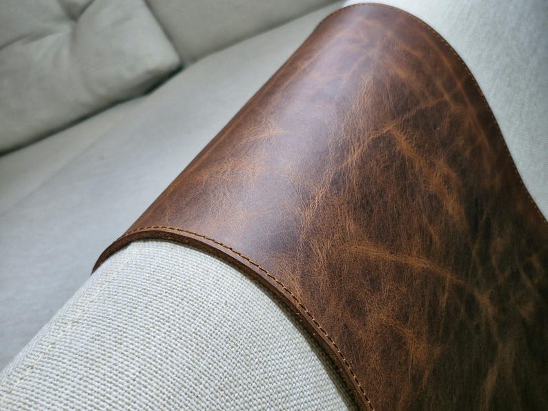 May include: A curved piece of brown leather with a visible grain and natural texture, featuring a smooth surface and a stitched edge. The leather is draped over a textured, light-colored fabric surface, highlighting the contrast in material.
