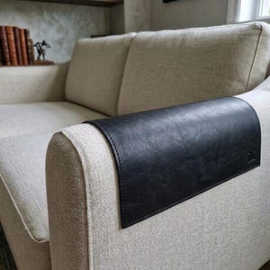 May include: A rectangular black leather armrest cover with visible stitching along the edges, draped over the arm of a beige fabric sofa. The cover features a small embossed logo on the lower right corner and has a smooth, textured surface.