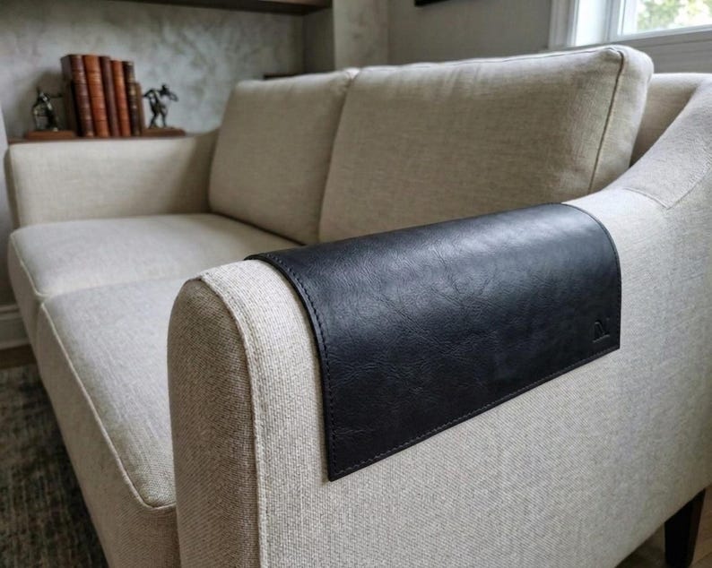 May include: A rectangular black leather armrest cover with visible stitching along the edges, draped over the arm of a beige fabric sofa. The cover features a small embossed logo on the lower right corner and has a smooth, textured surface.