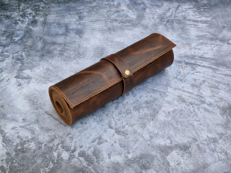 May include: A cylindrical brown leather roll-up case featuring a smooth texture and a single leather strap secured with a small brass-colored metal snap button fastener, designed for organized storage of various long items.