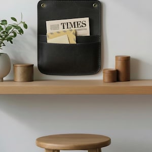 May include: A dark charcoal wall-mounted organizer featuring two pockets and two brass-toned grommets at the top. The organizer holds a newspaper with the word TIMES printed on it and a tan envelope. The item has visible stitching along the edges.