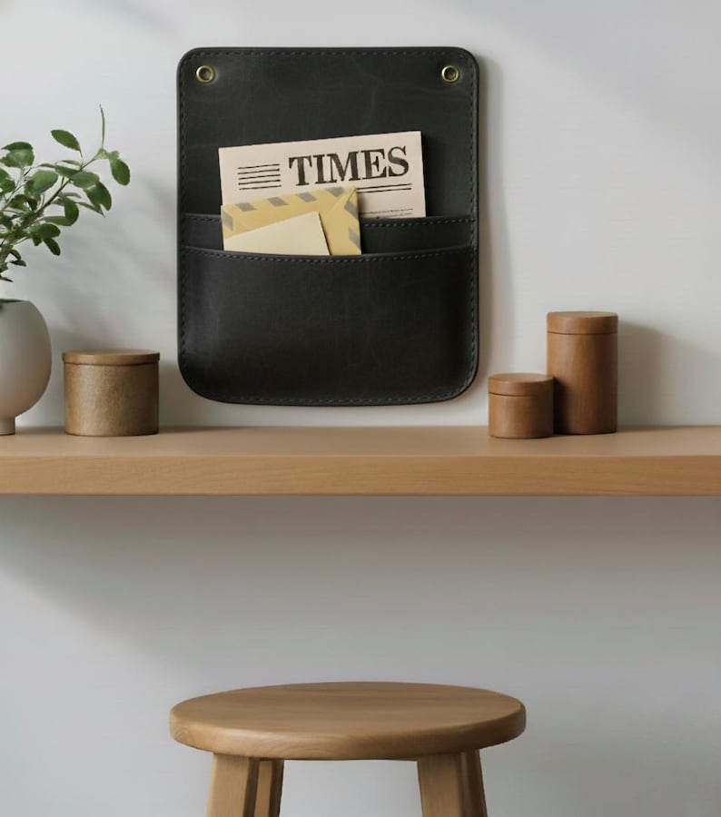 May include: A dark charcoal wall-mounted organizer featuring two pockets and two brass-toned grommets at the top. The organizer holds a newspaper with the word TIMES printed on it and a tan envelope. The item has visible stitching along the edges.