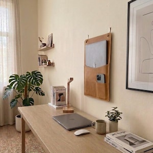 May include: A tan leather wall-mounted organizer featuring a large pocket for a laptop and a smaller front pocket for a smartphone. The organizer is suspended by two metal hooks on a light-colored wall above a wooden desk with a laptop and mouse.