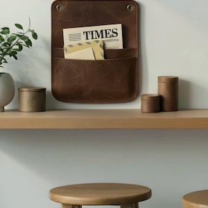 May include: A dark brown leather wall-mounted organizer with two pockets, featuring two brass-colored metal grommets at the top. The organizer holds a newspaper with the word TIMES printed on it and a tan envelope with a striped pattern.