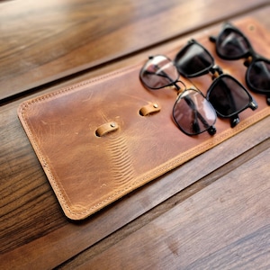 Puede incluir: Un soporte para gafas de cuero marr&oacute;n con tres pares de gafas de sol negras apoyadas en &eacute;l. El soporte tiene dos bucles cosidos para sujetar las gafas.