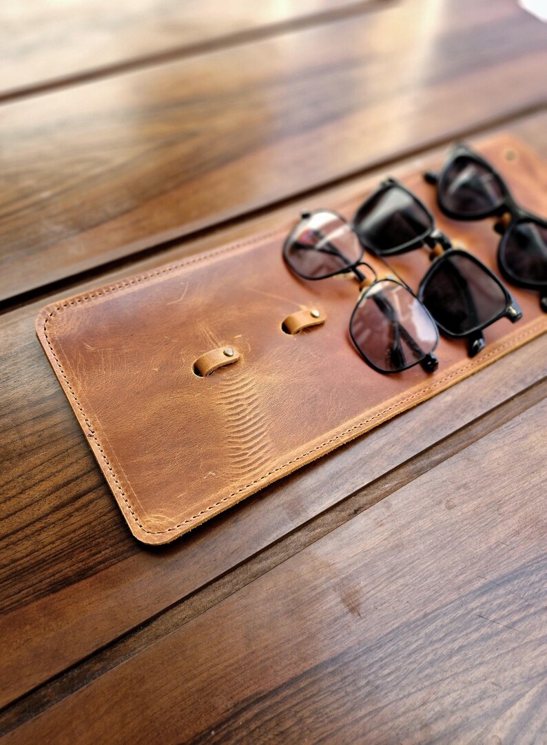 Puede incluir: Un soporte para gafas de cuero marr&oacute;n con tres pares de gafas de sol negras apoyadas en &eacute;l. El soporte tiene dos bucles cosidos para sujetar las gafas.
