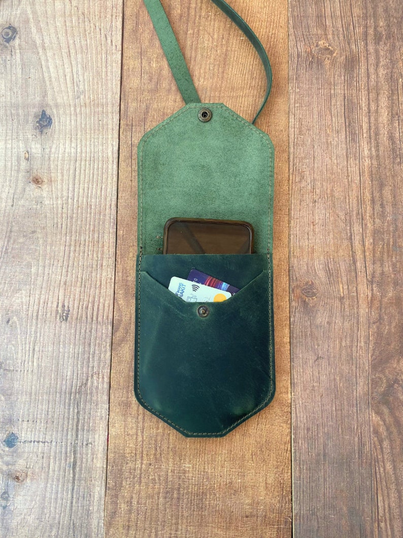 May include: A dark green leather phone case with a strap. The case has a flap that closes with a snap and a pocket for cards. A phone and cards are inside the case.