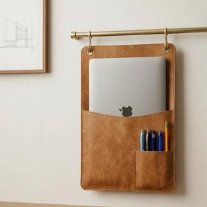 May include: A tan hanging wall organiser with a large pocket containing a silver laptop and a smaller front pocket holding four pens and a yellow pencil. The organiser is suspended from a gold-toned metal rod by two circular metal grommets.