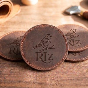 May include: A set of round, dark brown leather coasters featuring a debossed design of a bird perched on a branch above the letters RLZ. Each coaster has visible stitching along the circular edge, showcasing a smooth, matte finish and natural texture.