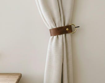 May include: A light beige curtain held back by a brown leather strap featuring a gold-toned metal ring and a central gold rivet. The strap wraps horizontally around the fabric, creating a gathered effect in the curtain material.