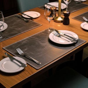 May include: A dining table set with gray leather placemats, white-rimmed plates, silverware, and wine glasses. The table is made of wood and has a warm, inviting tone, perfect for a restaurant or home setting.