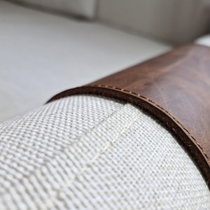 May include: A brown leather sleeve wrapped around a textured off-white fabric cylinder. The leather features visible stitching along the curved edge, showcasing a smooth, matte finish against the woven, light-colored textile surface.