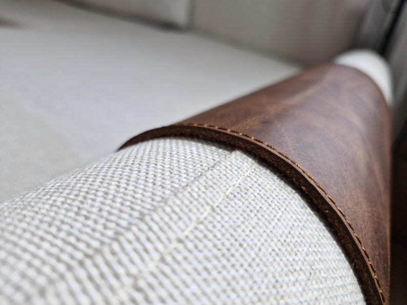 May include: A brown leather sleeve wrapped around a textured off-white fabric cylinder. The leather features visible stitching along the curved edge, showcasing a smooth, matte finish against the woven, light-colored textile surface.