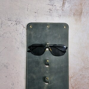 Puede incluir: Un estuche para gafas de cuero verde oscuro con tres ranuras para guardar gafas. Un par de gafas de sol negras con lentes oscuros descansa en la ranura superior.