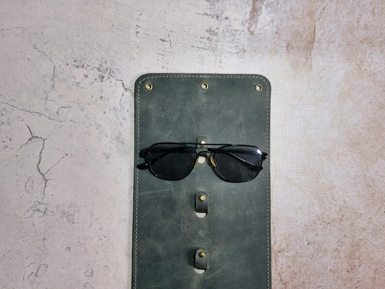 Puede incluir: Un estuche para gafas de cuero verde oscuro con tres ranuras para guardar gafas. Un par de gafas de sol negras con lentes oscuros descansa en la ranura superior.