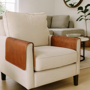 May include: A cream armchair with brown leather arm covers. The chair has a square back and armrests, with dark wooden legs. The leather arm covers are rectangular and cover the top of the armrests.
