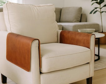 May include: An off-white upholstered armchair featuring two smooth, cognac-colored leather armrest covers draped over the sides. The chair has a thick back cushion and a seat cushion, supported by dark brown wooden legs on a light-colored floor.