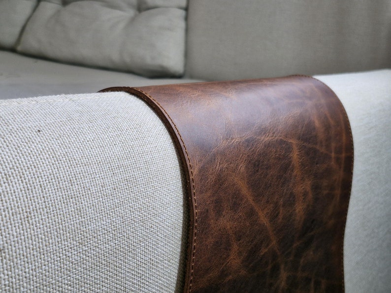 May include: A dark brown leather armrest cover with a smooth, distressed texture and visible stitching along the edges, draped over the light beige fabric arm of a sofa. The leather features natural grain patterns and a deep, rich color.