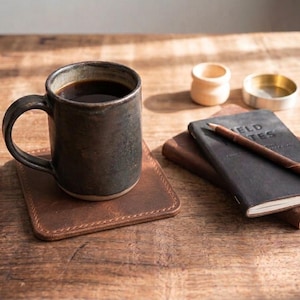 May include: A dark charcoal ceramic mug filled with black coffee sits on a square brown leather coaster. Nearby, a black Field Notes notebook with a brown pencil resting on top, a small light wood container, and a circular metal dish are visible.