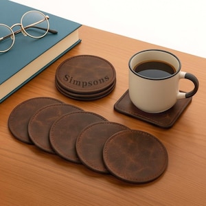 Puede incluir: Un conjunto de posavasos de cuero marrón, algunos redondos y otros cuadrados, con una taza de café y un libro. Los posavasos redondos están apilados y grabados con el nombre "Simpsons". La taza es blanca con borde y asa negros.