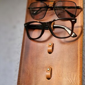 Puede incluir: Un soporte para gafas de cuero marr&oacute;n con dos ganchos de metal y dos detalles en lat&oacute;n. Dos pares de gafas cuelgan de los ganchos.