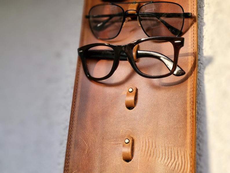 Puede incluir: Un soporte para gafas de cuero marr&oacute;n con dos ganchos de metal y dos detalles en lat&oacute;n. Dos pares de gafas cuelgan de los ganchos.