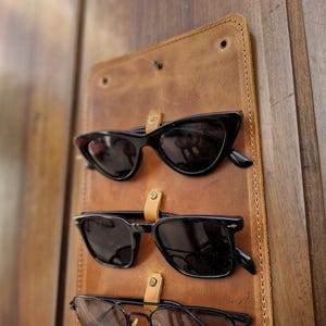 Puede incluir: Un soporte para gafas de sol de cuero marr&oacute;n con tres pares de gafas de sol negras colgadas.