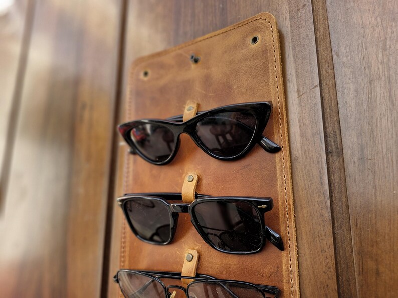 Puede incluir: Un soporte para gafas de sol de cuero marr&oacute;n con tres pares de gafas de sol negras colgadas.