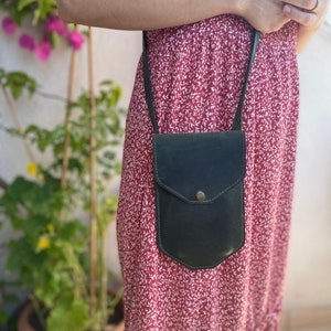 May include: A dark green leather crossbody bag with a flap closure and a small pocket on the front. The bag is worn by a person wearing a red floral dress.
