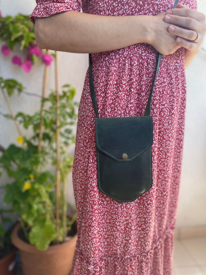 May include: A dark green leather crossbody bag with a flap closure and a small pocket on the front. The bag is worn by a person wearing a red floral dress.