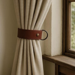 May include: Beige curtains held by a brown leather curtain tieback with a brass button and a metal ring. The curtains are hanging near a wooden window frame.