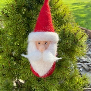 Könnte beinhalten: Ein Weihnachtsbaumschmuck aus Filz in Form eines Weihnachtsmanns. Der Schmuck ist rot und weiß mit einem weißen Bart und einer schwarzen Mütze.