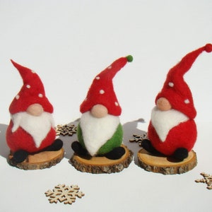 Peut inclure: Trois lutins de Noël en feutre avec des chapeaux rouges et blancs, des barbes blanches et des corps verts et rouges. Ils sont assis sur des tranches de bois.