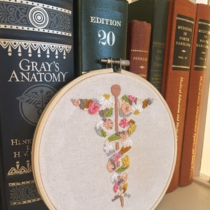 Puede incluir: Un primer plano de un aro de bordado blanco con un dise&ntilde;o floral colorido de un caduceo m&eacute;dico. El aro est&aacute; sentado en un estante con varios libros detr&aacute;s. Los libros tienen t&iacute;tulos como "Gray's Anatomy" y "Medicine in North Carolina".