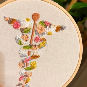 Puede incluir: Un aro de bordado blanco con un dise&ntilde;o floral en forma de caduceo m&eacute;dico. Las flores son en tonos de rosa, blanco, amarillo, verde y marr&oacute;n.