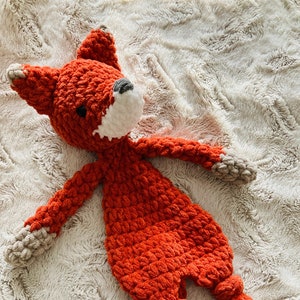 May include: A crocheted orange fox plush toy with white and gray accents on the tail. The toy is lying on a white fluffy surface.