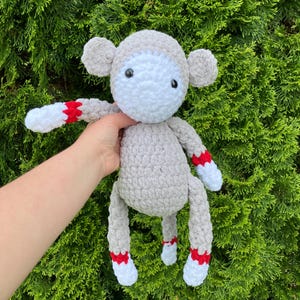 May include: A handmade crochet sock monkey doll in shades of gray, white, and red. The monkey has a gray body, white face and hands, and red and white accents on the arms and legs. The doll is held against a backdrop of green foliage.
