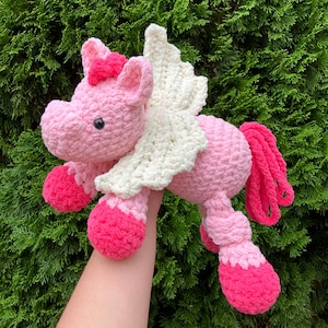 May include: A handmade, pink crochet unicorn with cream-colored wings and a bright pink mane and hooves. The plush toy has a sweet expression and is designed with a soft, textured yarn. A charming, whimsical creation.