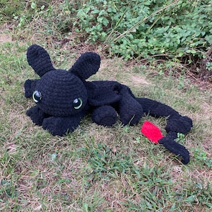 May include: A black crocheted dragon plush toy with large, round green eyes and a red fin. The dragon is lying on green grass, with its tail extended. The plush has a soft, textured appearance.