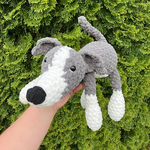 May include: A handmade crochet dog toy in gray and white. The dog has a long snout, floppy ears, and a black nose. The toy is soft and plush, with a textured appearance. The dog's body is gray, with white accents on the face and paws.
