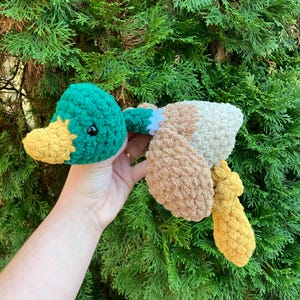 May include: A handmade crochet duck toy with a green head, yellow beak, and brown and cream-colored body. The duck has a fluffy texture and black eyes. The toy is held against a backdrop of green foliage.