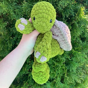 May include: A handmade, plush turtle in shades of green and tan. The turtle has a textured, fuzzy appearance, with a green body and limbs, and a tan shell. The turtle's feet have tan accents. The eyes are small and black.
