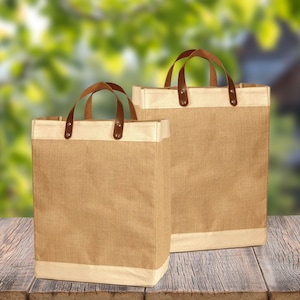 May include: Two brown burlap tote bags with leather handles and a white trim. The bags are sitting on a wooden surface.