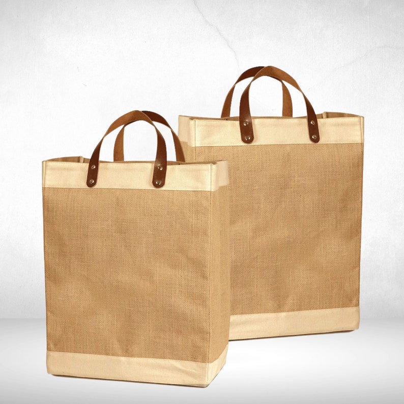 May include: Two brown burlap tote bags with leather handles and a white trim. The bags are empty and sitting on a white surface.