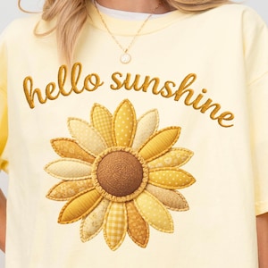 Op de afbeelding: Een lichtgeel t-shirt met de geborduurde tekst "hello sunshine" boven een groot zonnebloemontwerp. De zonnebloem heeft verschillende gele patronen en een bruin midden. Het shirt is een casual, comfortabele stijl.