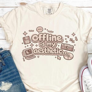 Puede incluir: Camiseta color crema con el texto "Offline is my aesthetic" en marrón. El diseño incluye ilustraciones de un disquete, un portátil, una cámara y un CD. La camiseta se combina con pantalones cortos vaqueros y zapatillas blancas.